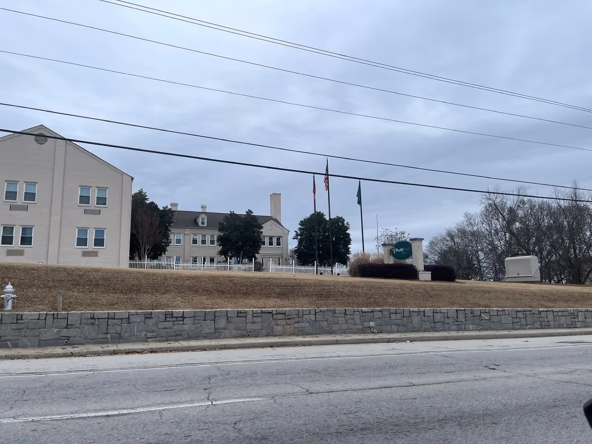Exterior view of a beige multi-story senior living building on a grassy hill with flagpoles and a Pruitt sign, seen from the road.