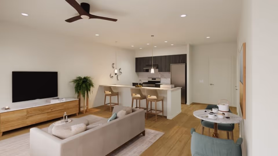 A modern open-concept living space in a senior residence featuring a beige sofa with cushions, a wooden TV stand with a flat-screen TV, a potted plant, and a ceiling fan. The kitchen area has dark cabinets, a stainless steel refrigerator, an oven, and a white countertop with three bar stools. A small round dining table with two teal chairs is set with a teapot and cups. The room has wooden flooring and recessed lighting.