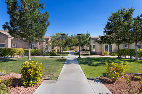 Paved walkway through a landscaped courtyard flanked by single-story cottage-style buildings and trees under a clear blue sky.