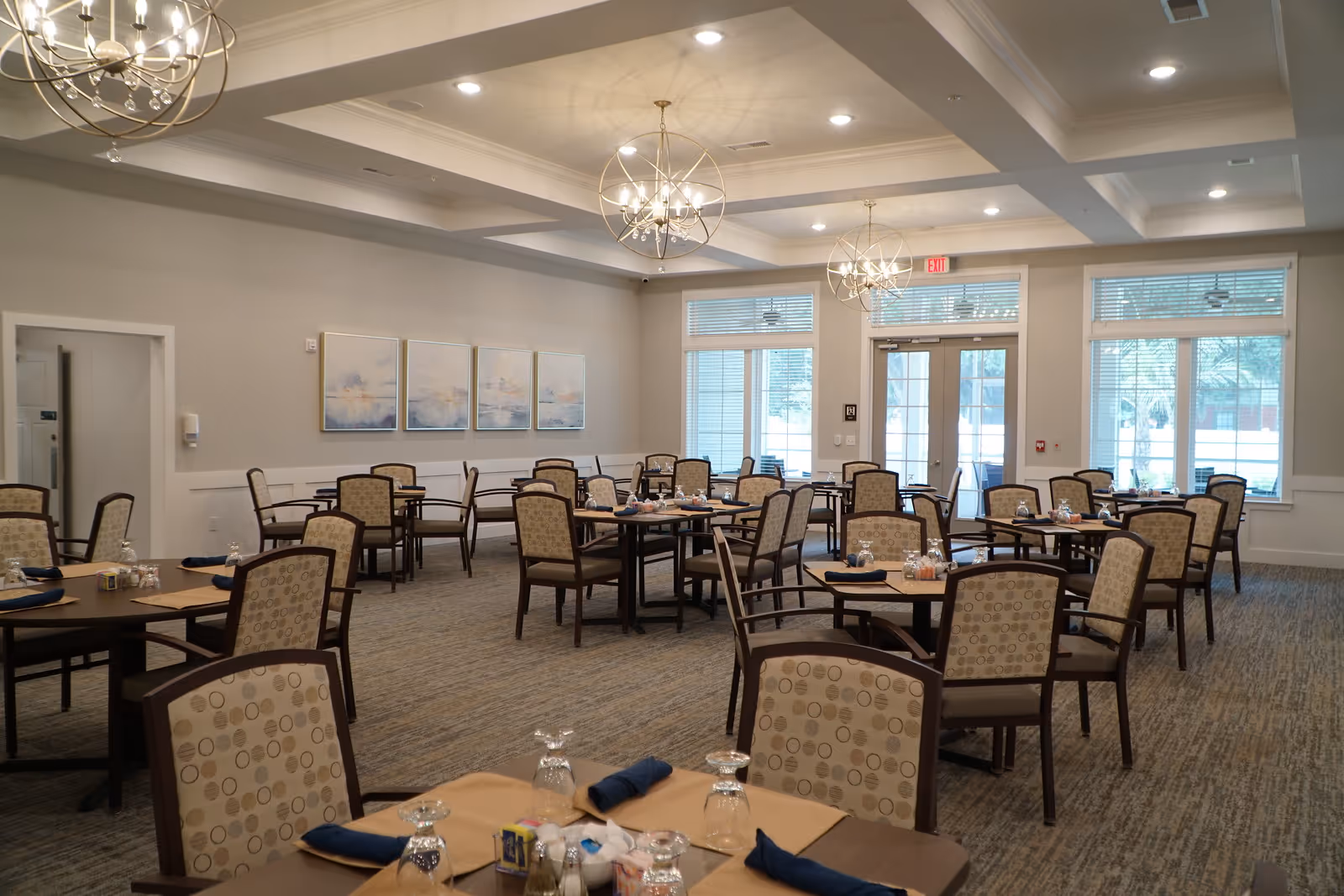 Spacious dining room with multiple set tables and cushioned chairs, chandeliers, and large windows letting in natural light.