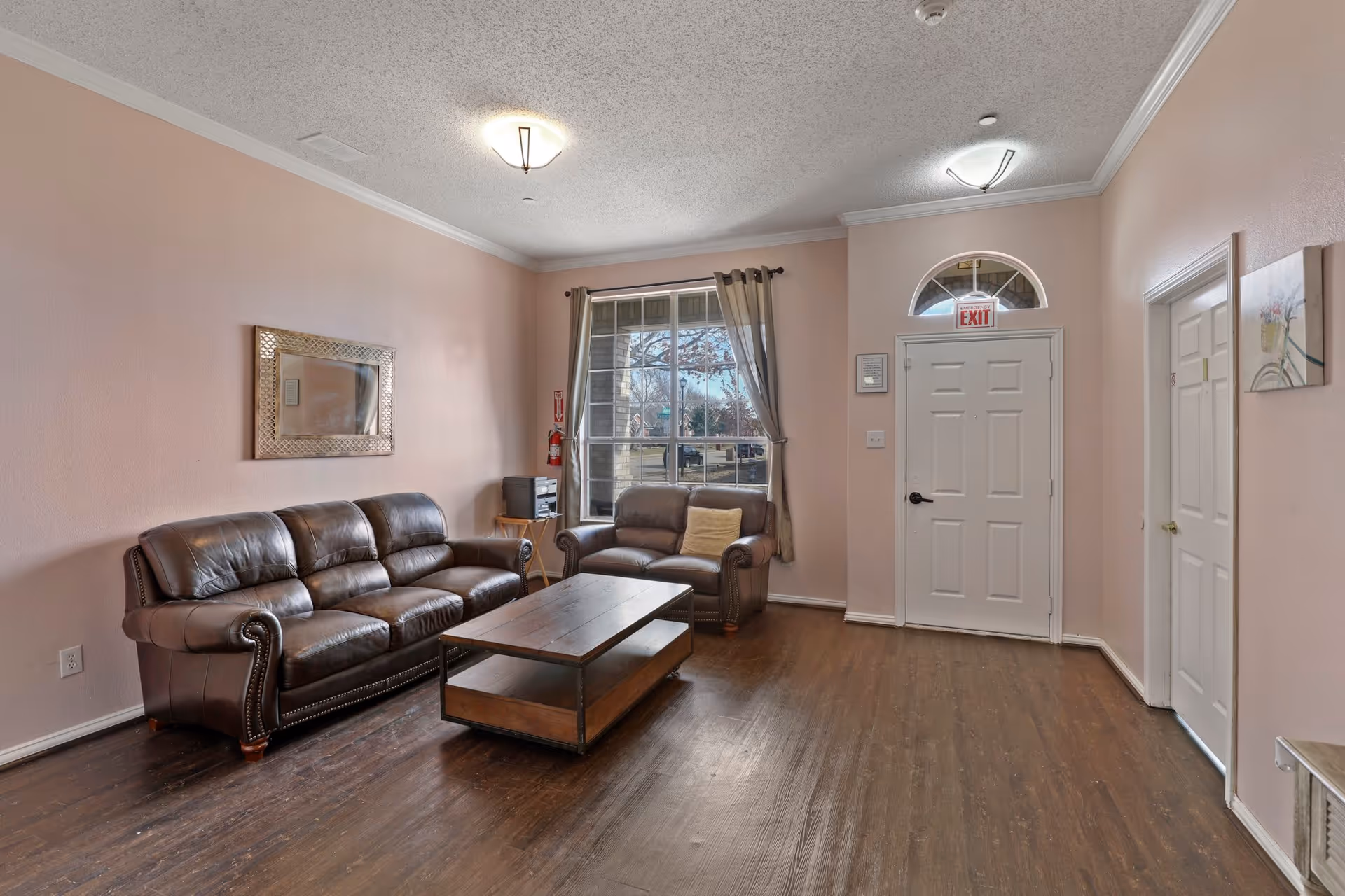 A cozy living room with light pink walls, dark wooden flooring, a brown leather sofa and loveseat, a wooden coffee table, a large window with curtains, an exit door with an exit sign above it, and a wall mirror above the sofa.