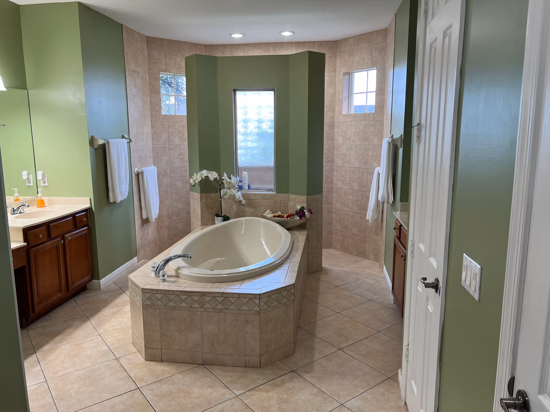 A spacious bathroom with a large built-in bathtub in the center surrounded by beige tiles. The walls are painted green with two towel racks holding white towels. There are two wooden vanities with sinks and mirrors on either side of the room. Two small frosted windows allow natural light to enter.