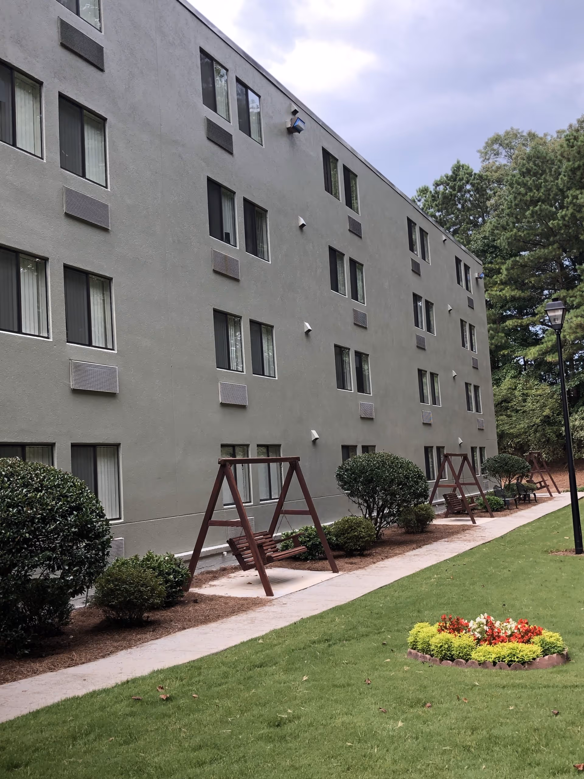 Exterior view of a multi-story senior housing building with a walkway, wooden swing benches, shrubs, and a small flowerbed on a grassy lawn.