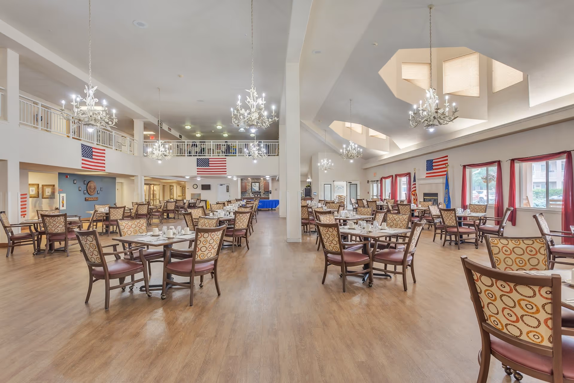 Spacious dining room in a senior living facility with multiple tables and chairs arranged neatly. The room features wooden flooring, chandeliers hanging from the ceiling, large windows with red curtains, and American flags displayed on the walls. The area is well-lit with natural and artificial light.