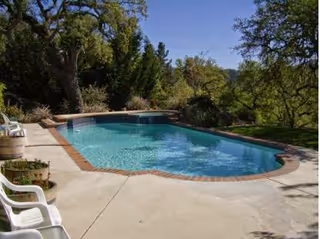 Outdoor swimming pool surrounded by concrete deck with white plastic chairs and trees in the background under a clear blue sky.
