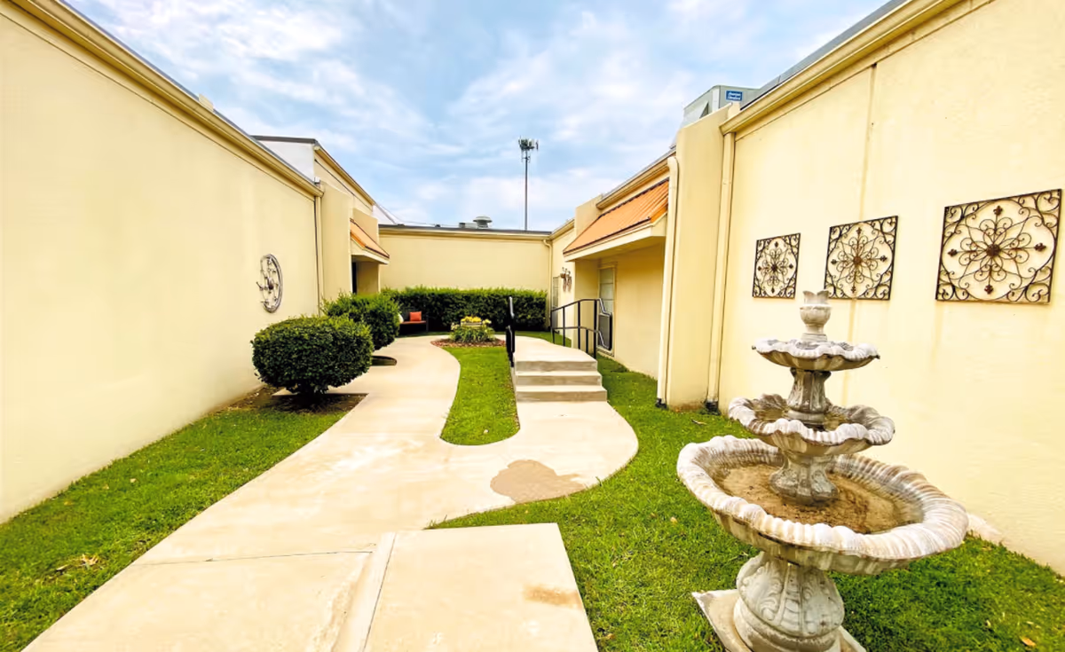 Outdoor courtyard with a curved concrete walkway, a three-tier fountain, trimmed shrubs and beige building walls with decorative wall art.