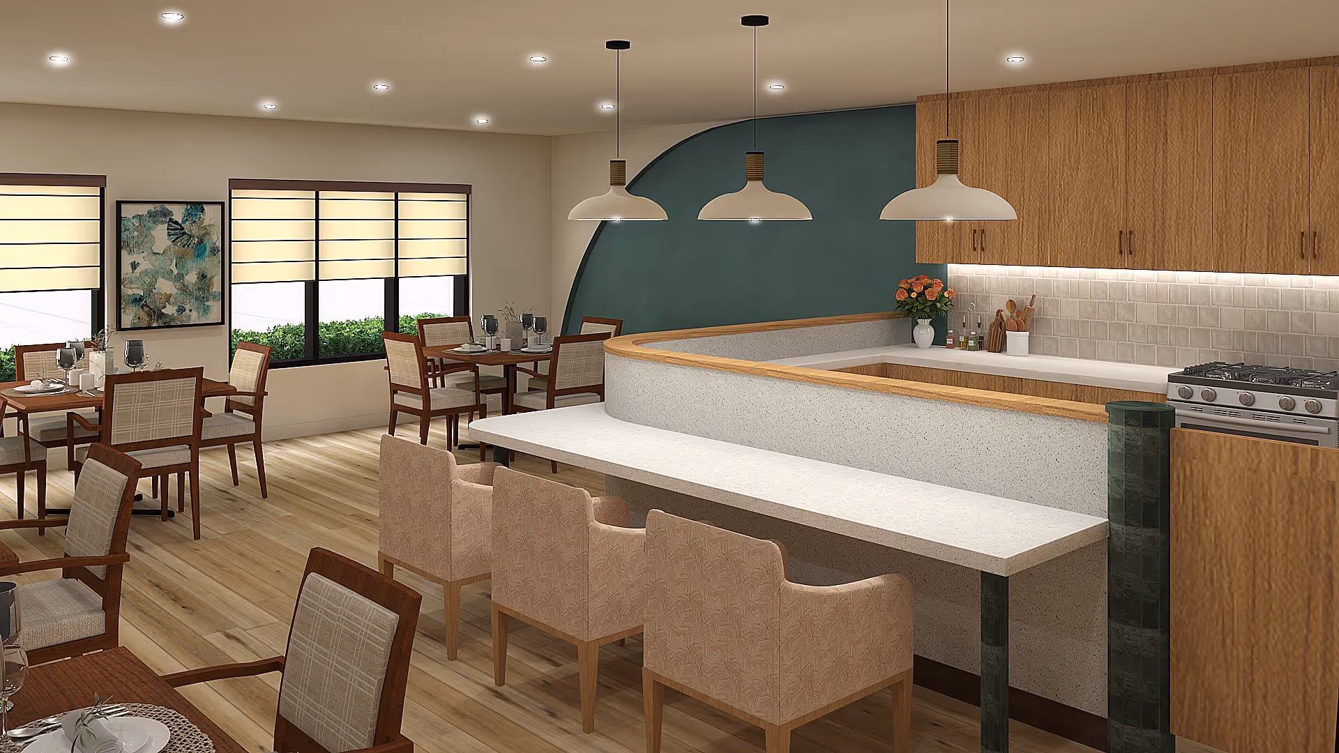 Interior view of a dining area and kitchen in a senior living facility. The dining area has multiple wooden tables with chairs set for meals, featuring placemats, plates, and glasses. The kitchen area includes a countertop with three pendant lights hanging above, wooden cabinets, a stove, and decorative items such as a vase with flowers and kitchen utensils. The room has light wood flooring, large windows with beige blinds, and a teal accent wall.