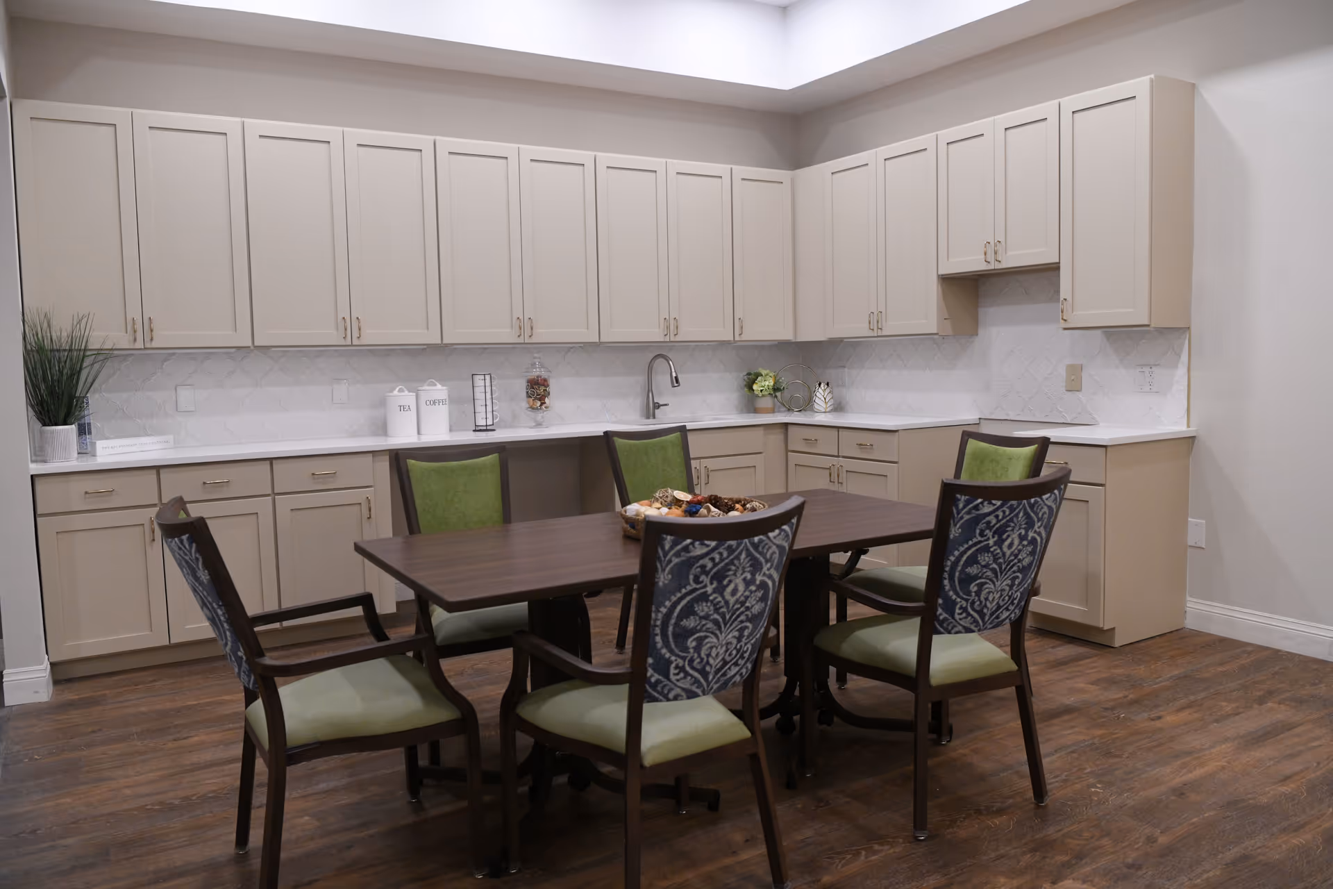 A modern kitchen area with beige cabinets and white countertops arranged in an L-shape. There is a dark wooden table with six chairs around it, featuring green seats and patterned backs. The floor is wooden, and the walls are painted white. Decorative items including a plant, canisters labeled Tea and Coffee, and a bowl with decorative balls are placed on the countertop and table.