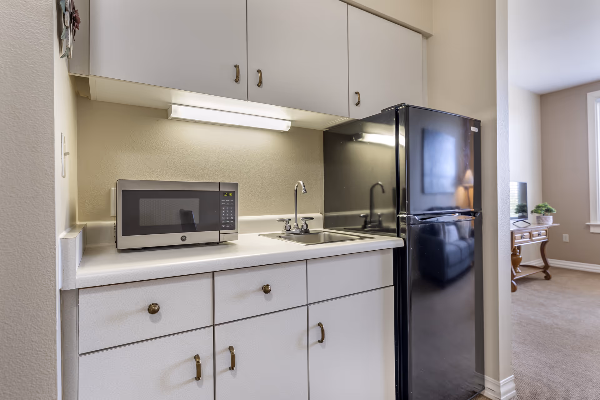 Small kitchenette with white cabinets, a microwave, sink and a black refrigerator opening into a living area.