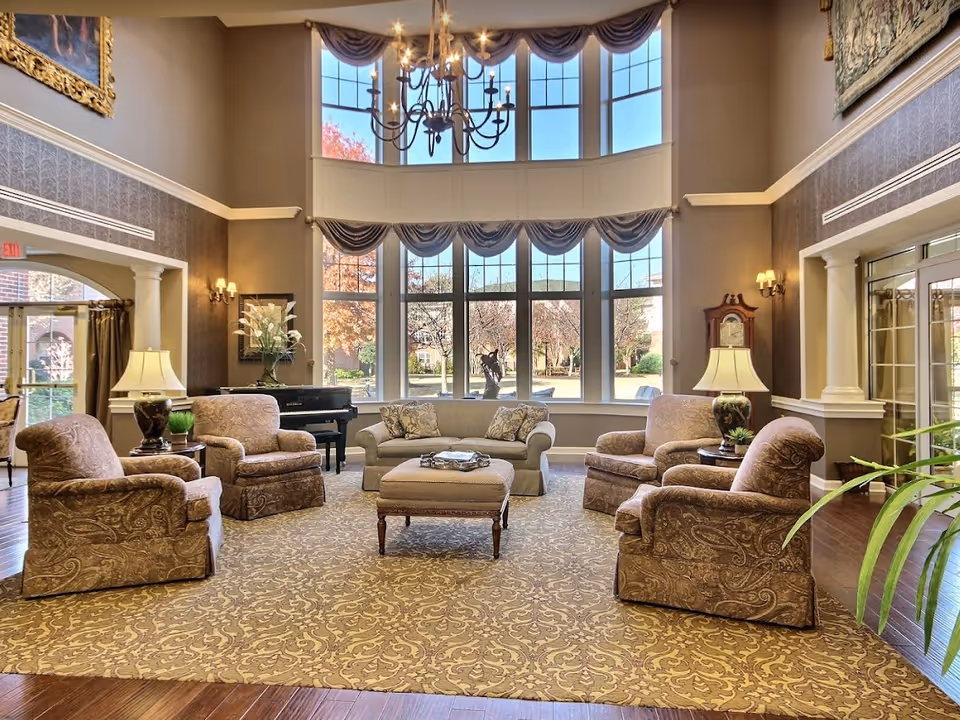 A spacious and elegant living room with high ceilings and large windows letting in natural light. The room features a chandelier, a patterned carpet, four upholstered armchairs arranged around a central ottoman, a sofa with decorative pillows, two table lamps on side tables, a grand piano, and a grandfather clock. Outside the windows, trees with autumn foliage are visible.