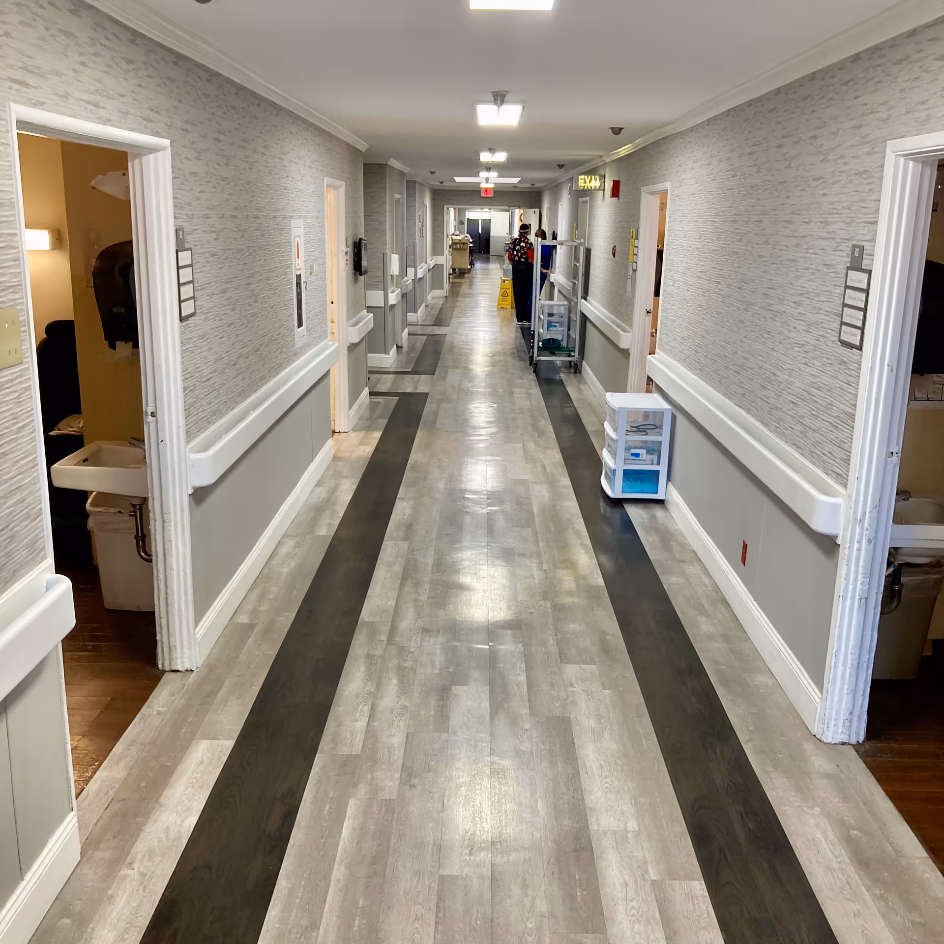 Long hallway in a senior living facility with light gray walls and wood-patterned flooring featuring dark stripes. Several open doors on both sides reveal rooms with sinks and some furniture. A cleaning cart and a caution wet floor sign are visible in the distance. Ceiling lights illuminate the corridor.