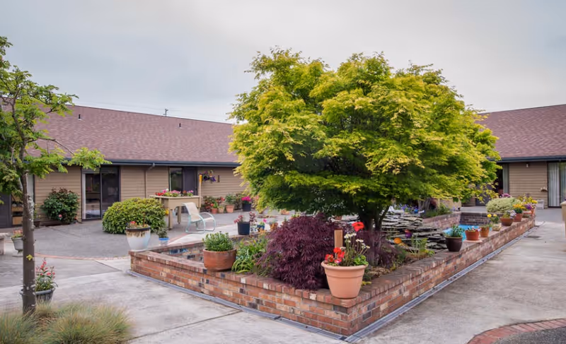 A landscaped courtyard with a raised brick planter, potted flowers and a small tree in front of a single-story senior living building.