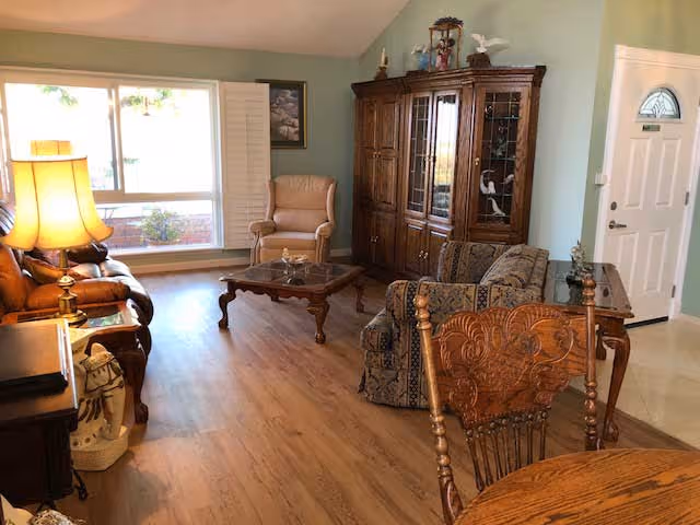 A cozy living room with wooden flooring, a large window with white shutters, a brown leather sofa, a beige armchair, a patterned armchair, a wooden coffee table, a wooden china cabinet with glass doors, and a wooden dining table with carved chairs. A lamp is lit on a side table next to the sofa, and the front door is visible in the background.