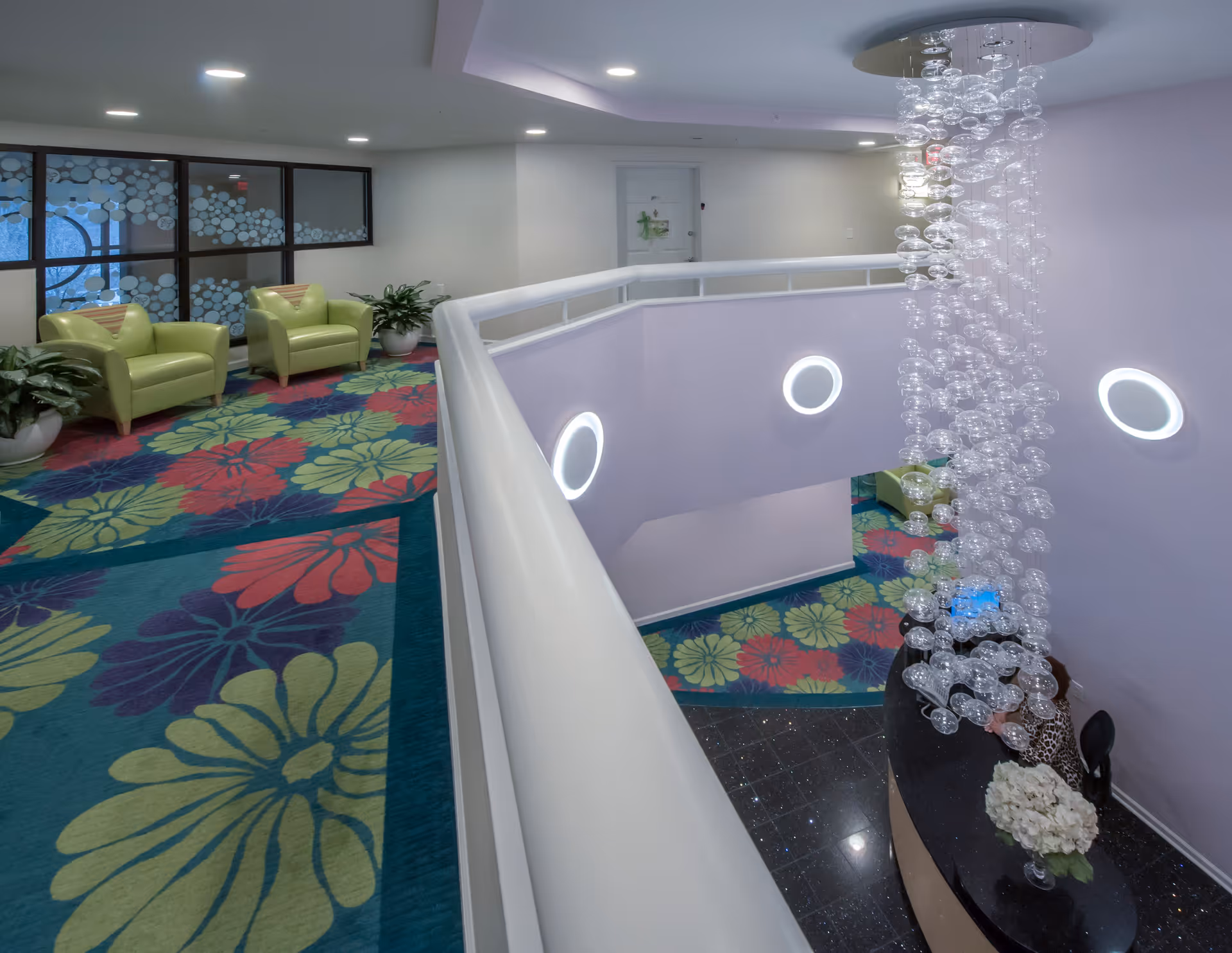 Interior view of a senior living facility featuring a colorful floral carpet, two green armchairs, potted plants, a white railing overlooking a lower level with a modern chandelier made of hanging glass spheres, and a reception desk with a flower arrangement.