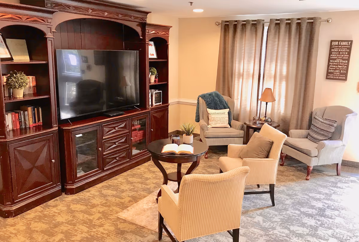 A cozy living room with a large wooden entertainment center holding a flat-screen TV. There are four armchairs arranged around a small round coffee table with an open book and a small plant on it. The room has beige walls, a window with closed curtains, a side table with a lamp, and a decorative wall sign.