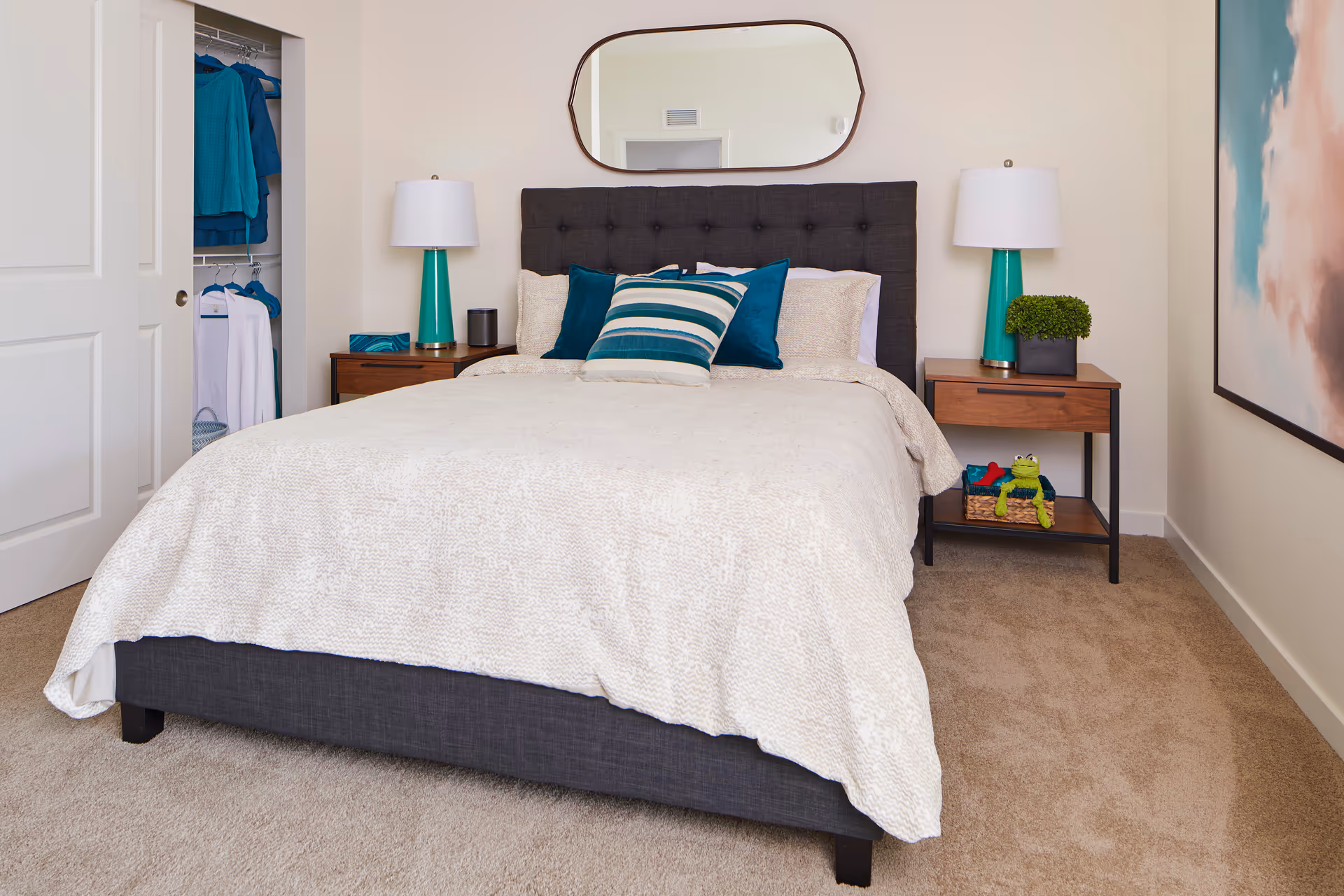 A neatly made bed with a dark upholstered headboard and cream-colored bedding in a bedroom. There are three decorative pillows in shades of teal and cream on the bed. On each side of the bed are wooden nightstands with teal lamps, a small plant, and a basket with a frog plush toy. A closet with clothes hanging is visible on the left side, and a large abstract painting is on the right wall. A mirror hangs above the headboard.
