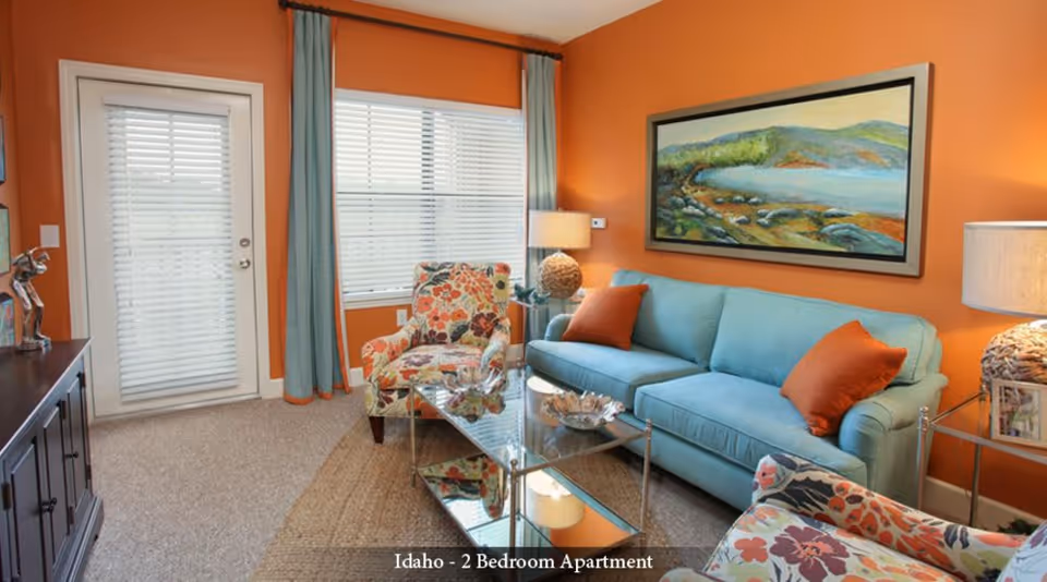 A cozy living room with orange walls, a light blue sofa with orange pillows, two floral patterned armchairs, a glass coffee table, a side table with a lamp, and a large landscape painting on the wall. There is a window with blinds and blue curtains, and a door with blinds leading outside.