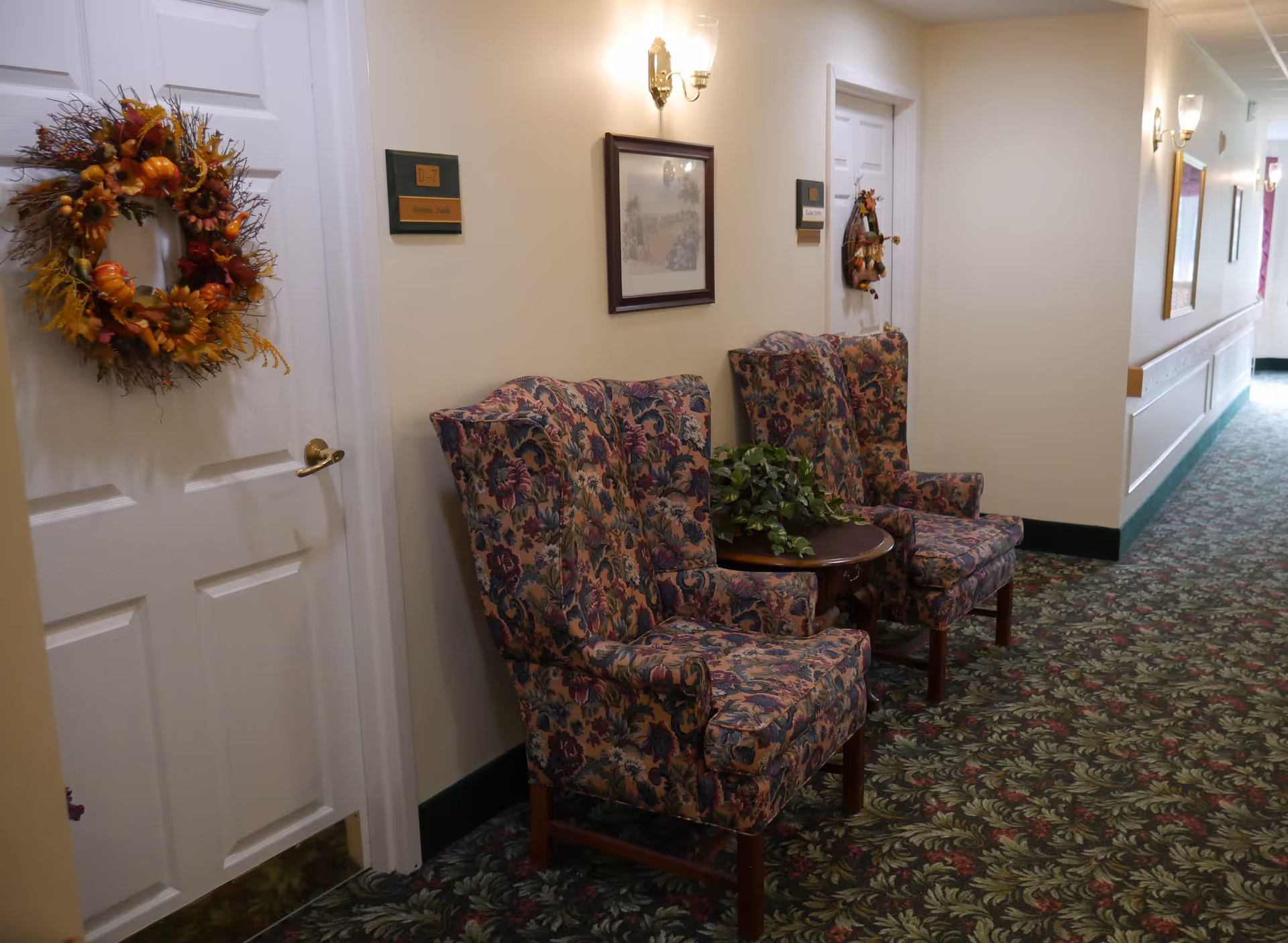 Carpeted hallway with two floral upholstered armchairs and a small table between doors decorated with autumn wreaths.