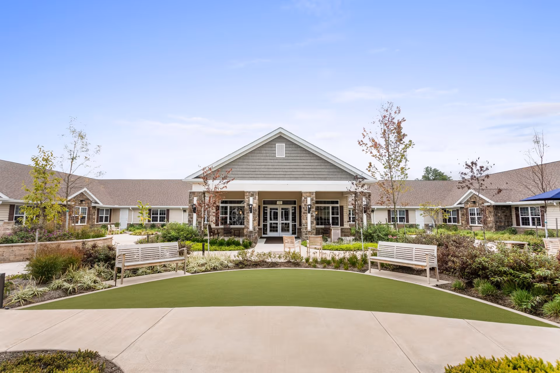 Front exterior view of a single-story senior living facility with a covered entrance supported by stone pillars, surrounded by landscaped gardens, benches, and a clear blue sky.
