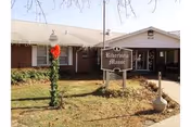 Exterior view of a single-story brick building with a sign reading 'Riverside Manor' in front. The building has a covered entrance and a small lawn with a decorated lamp post and a large ceramic pot near the sidewalk.
