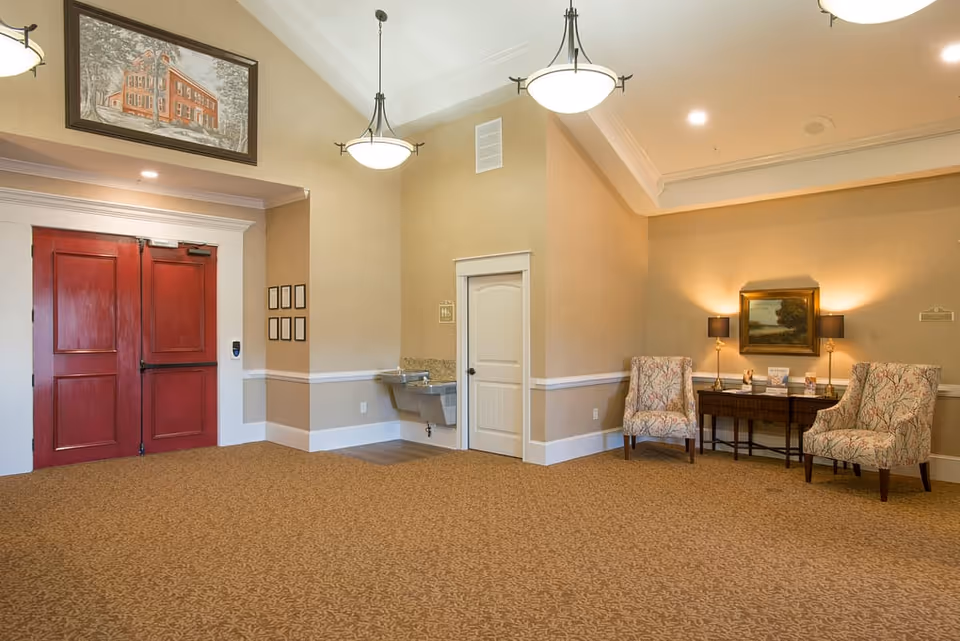 Spacious reception lounge with red double doors, a seating area of two patterned armchairs and a wooden side table beneath a landscape painting, pendant lights, and a drinking fountain.
