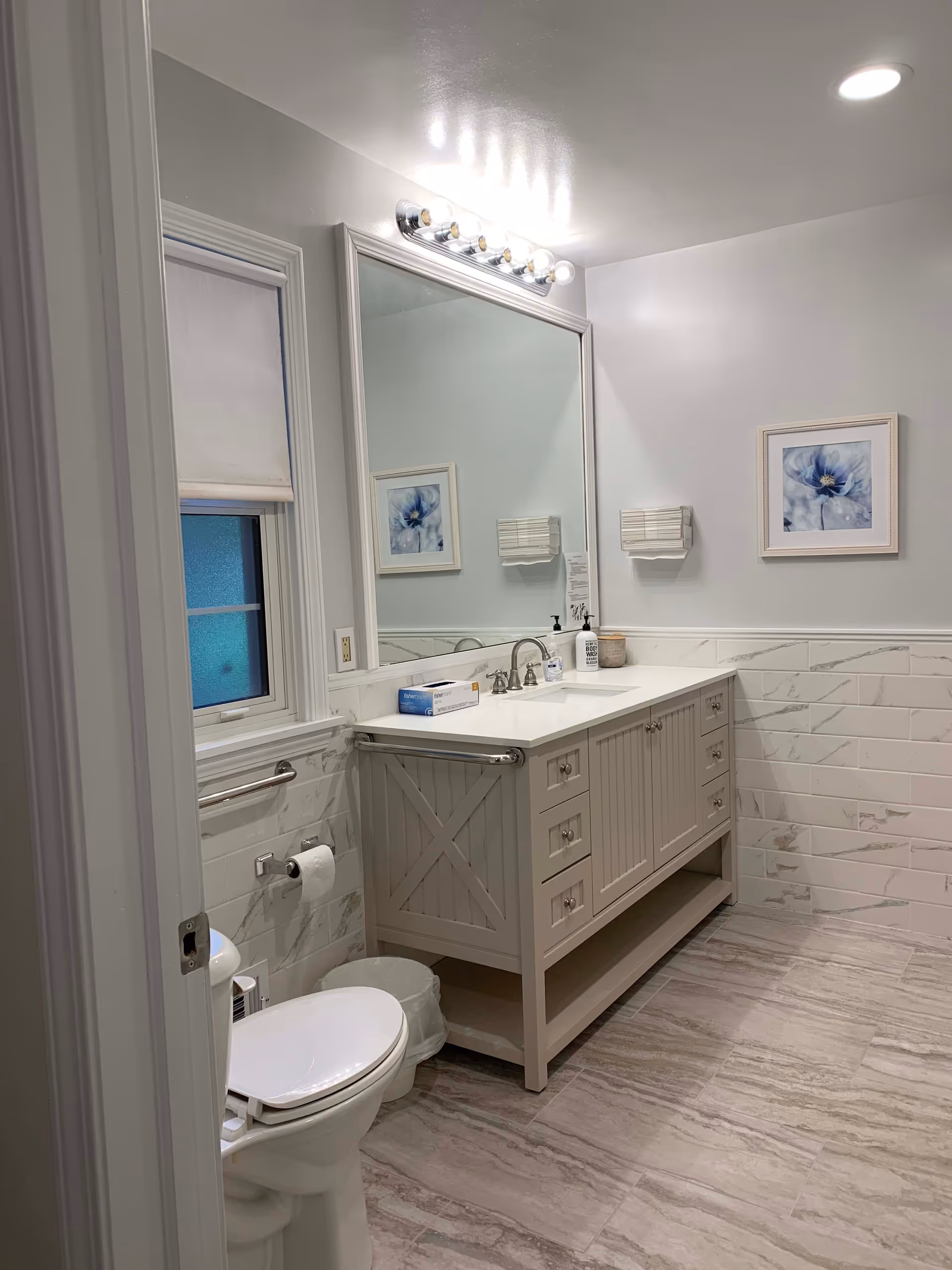 A clean and modern bathroom with a white toilet, a large vanity with a white countertop and sink, a large mirror above the vanity, a window with a white blind, a wall-mounted light fixture with multiple bulbs, and a framed floral picture on the wall. The floor and lower walls are tiled in a light gray marble pattern.