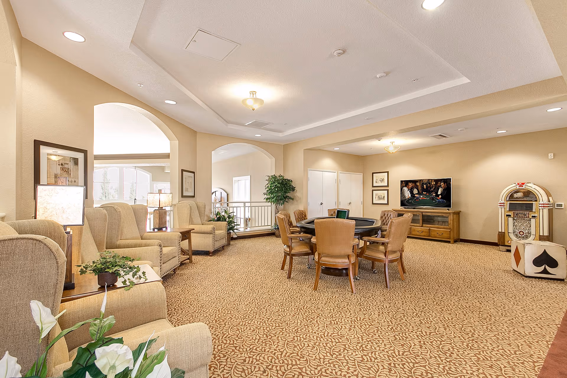 A spacious, well-lit common area in a senior living facility featuring beige upholstered armchairs arranged along the left wall with side tables and lamps. In the center, there is a round table surrounded by wooden chairs. On the right side, a flat-screen TV is mounted on a wooden cabinet, and a vintage jukebox stands next to it. The room has beige walls, patterned carpet, and recessed lighting with a ceiling light fixture.