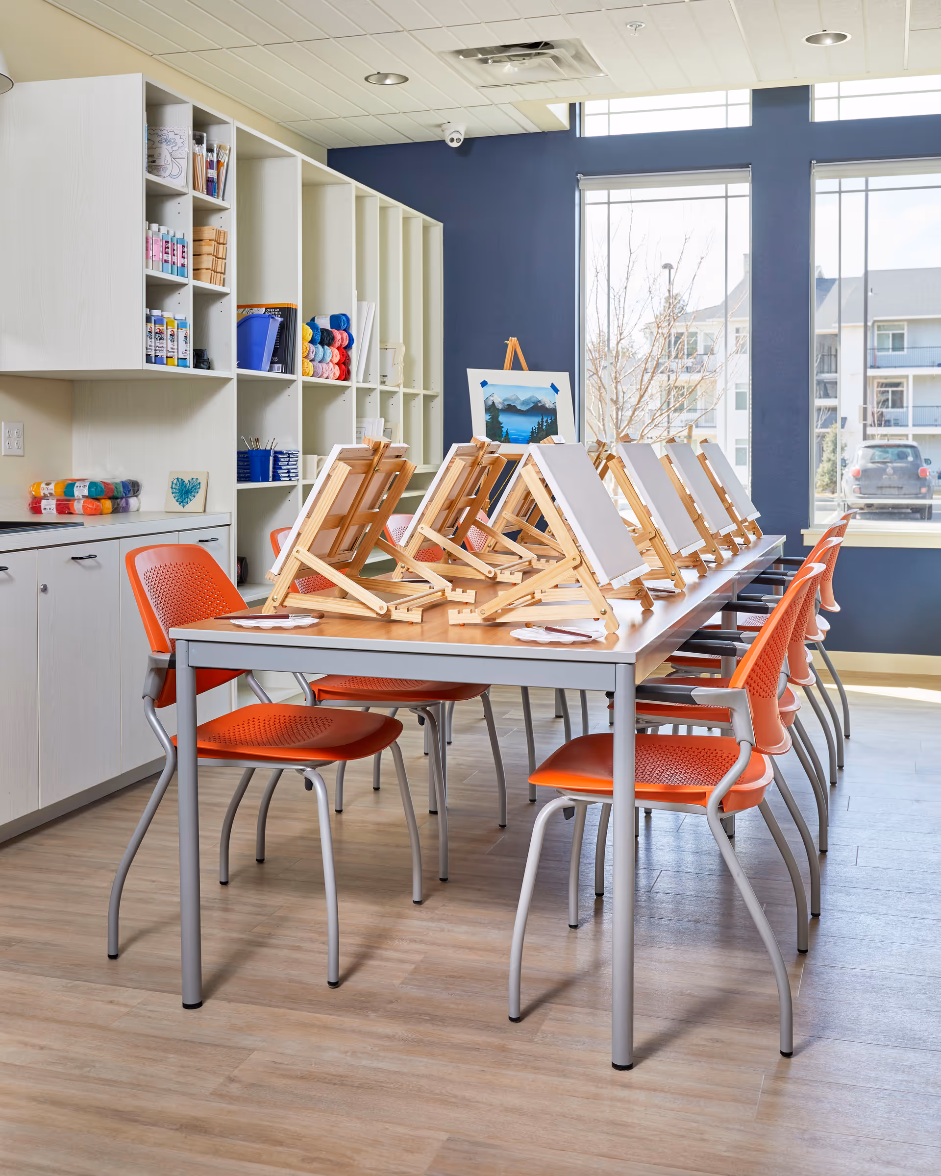 A bright art room with a long table set up with wooden easels holding blank canvases. Orange chairs are arranged around the table. Shelves on the left wall hold various art supplies including paint bottles, brushes, and yarn. Large windows on the right side let in natural light and show a view of buildings and parked cars outside.