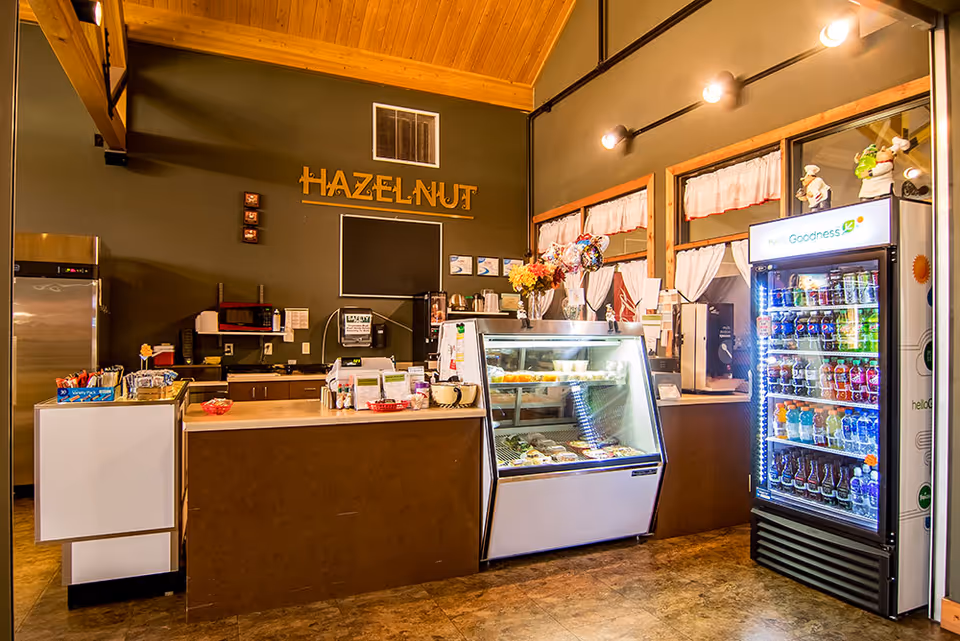 Interior view of a small cafe or snack bar area named 'Hazelnut' with a counter, refrigerated display case with food items, a beverage cooler stocked with drinks, and a kitchen area in the background. The space has warm lighting and wooden ceiling beams.