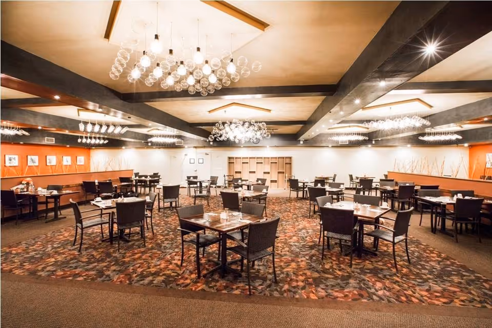 A spacious dining room with multiple square wooden tables and dark chairs arranged neatly on a patterned carpet. The room features modern chandeliers with multiple glass bulbs hanging from the ceiling, warm lighting, and orange accent walls with framed artwork and decorative branches.