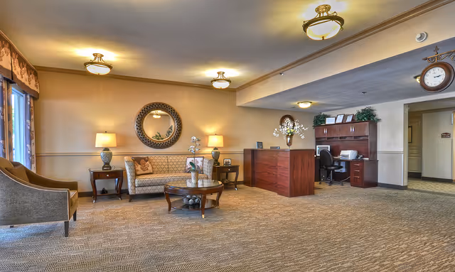 A spacious and well-lit reception area with a wooden front desk, a seating area including a sofa, armchair, and coffee table, decorative lamps, a round wall mirror, and a clock on the wall. The room has beige walls and carpeted flooring.