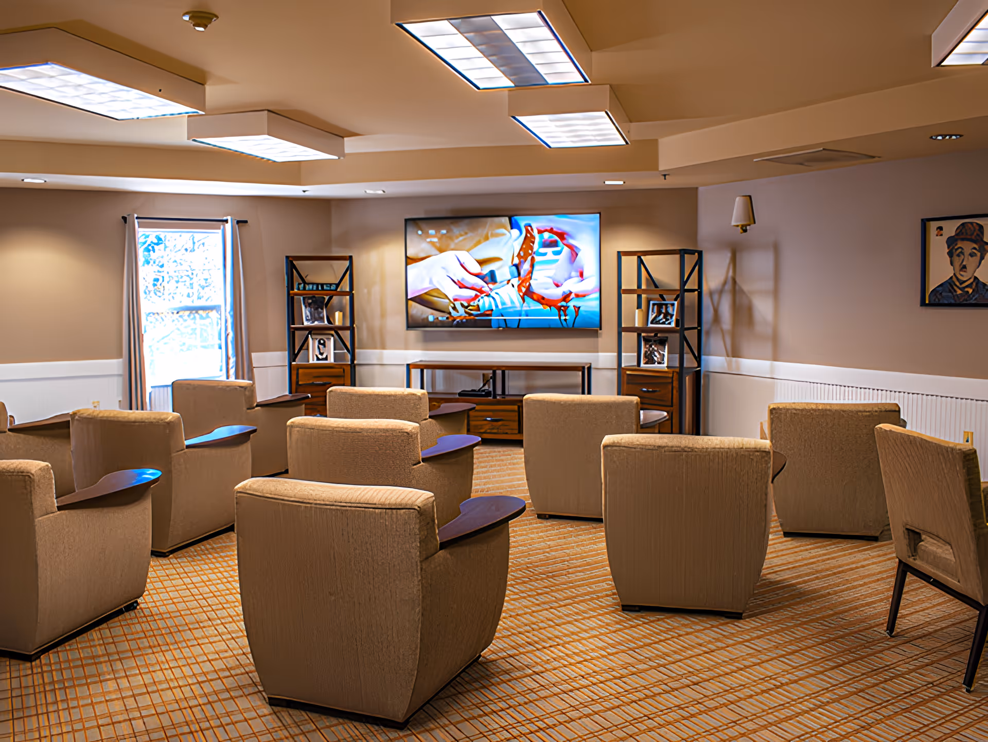 A cozy common room with multiple beige armchairs arranged in rows facing a large flat-screen TV mounted on the wall. The room has beige walls with white wainscoting, a window with curtains on the left, two wooden shelving units with framed photos and decorative items flanking the TV, and a patterned carpeted floor. The ceiling has several rectangular fluorescent light fixtures.
