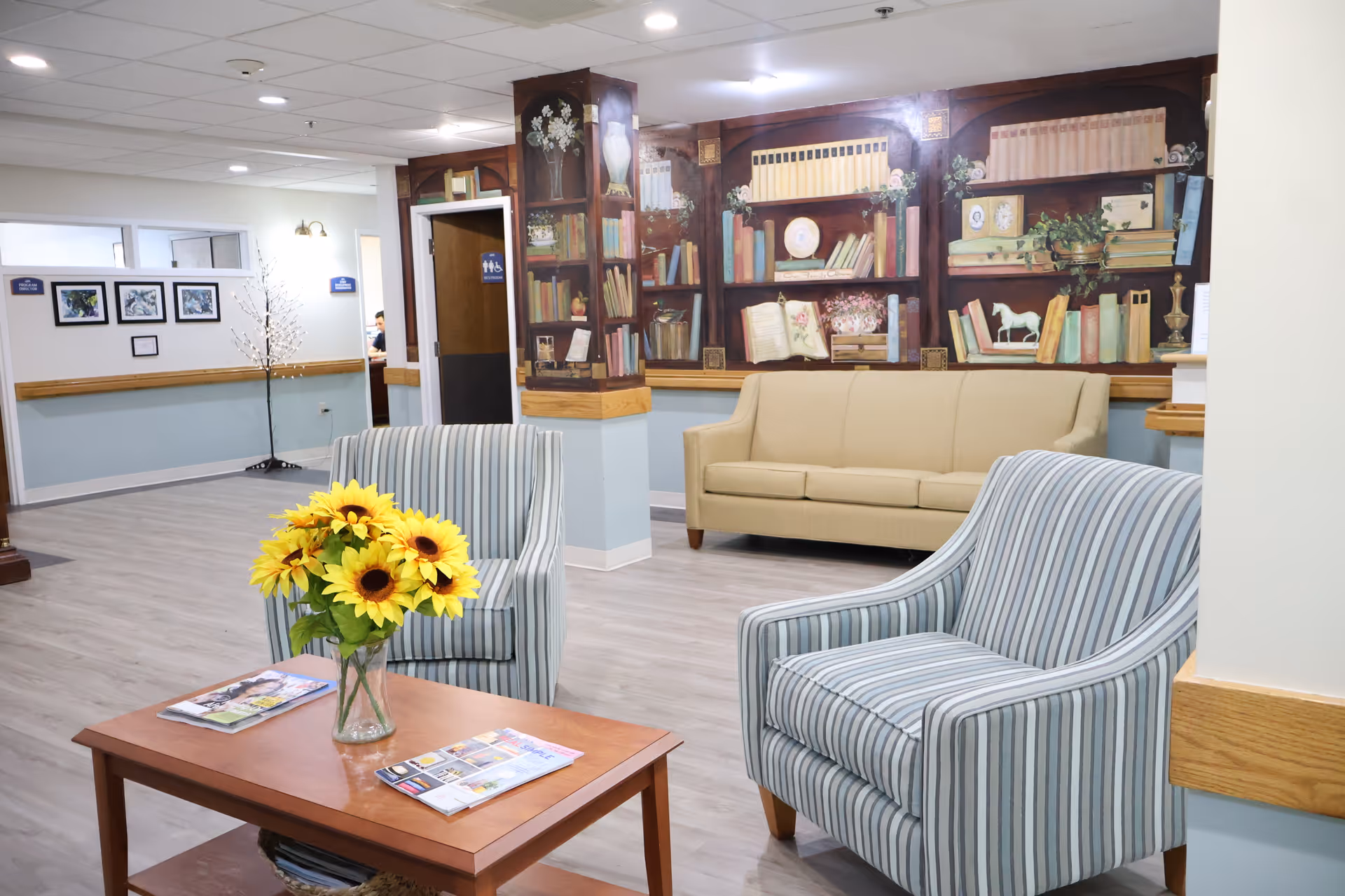 A cozy seating area in a senior living facility with two striped armchairs and a beige sofa. A wooden coffee table with a vase of sunflowers and magazines is in the center. The background features a wall mural designed to look like bookshelves filled with books and decorative items. The floor is light wood, and the walls are painted in soft blue and white tones. A hallway with framed pictures and a restroom door is visible in the background.