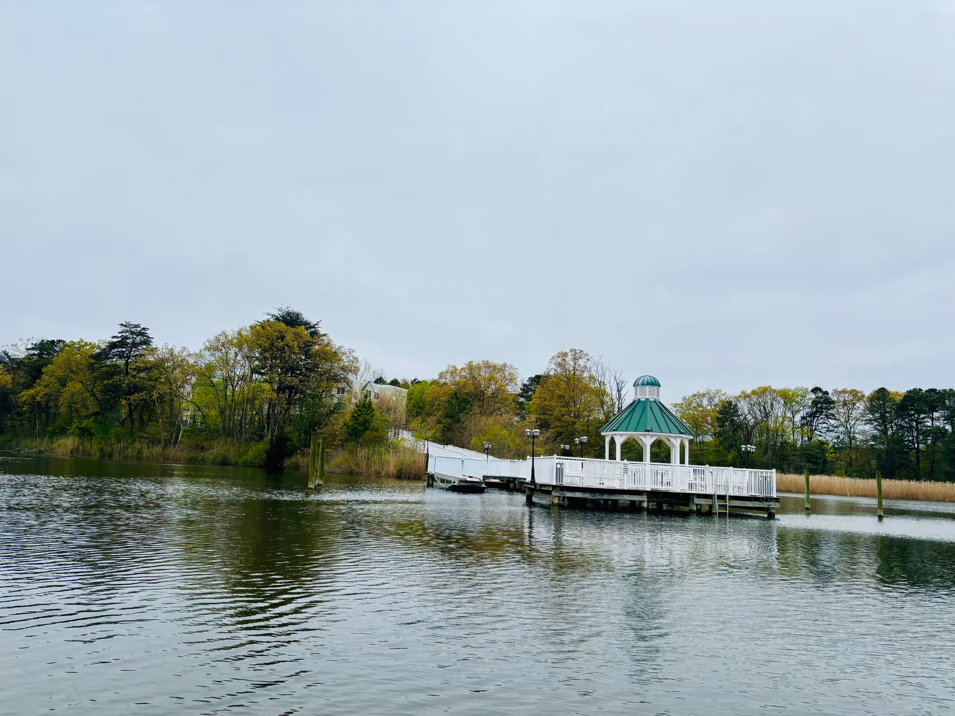 A white gazebo with a green roof situated on a wooden pier extending over a calm body of water, surrounded by trees with green and yellow foliage under a cloudy sky.