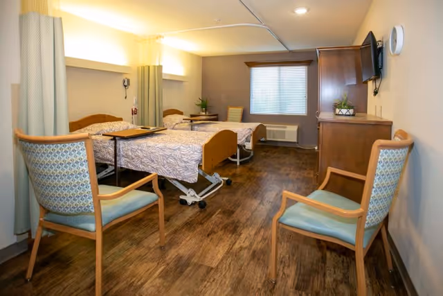 Shared healthcare room with two adjustable beds, bedside tables, chairs, a wall-mounted TV, and wood-look flooring.