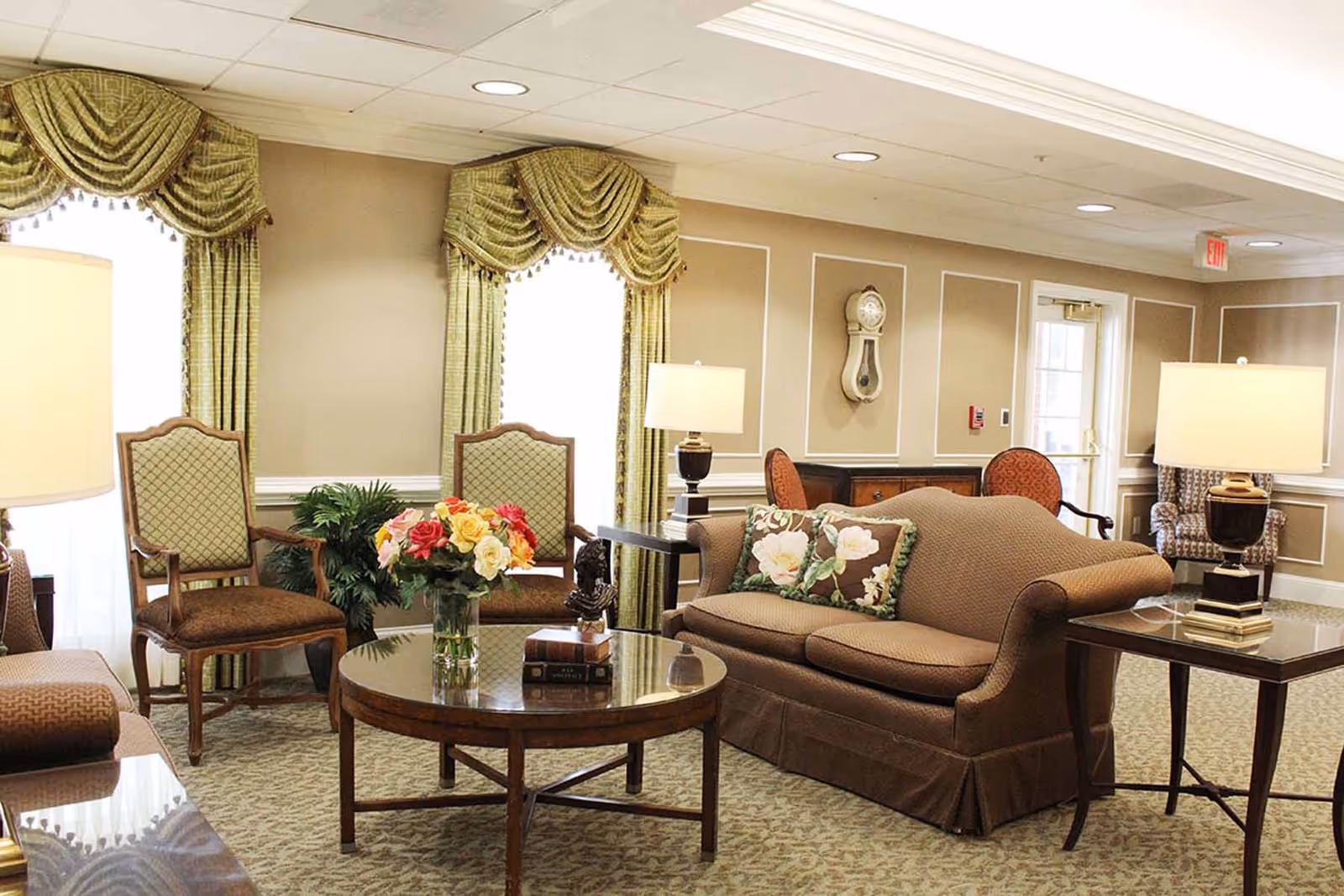 A cozy living room area with a brown upholstered sofa adorned with floral pillows, two wooden armchairs with patterned cushions, a round wooden coffee table with a vase of colorful flowers and decorative items, two side tables with lamps, green draped curtains on windows, and a wall clock mounted on beige paneled walls.