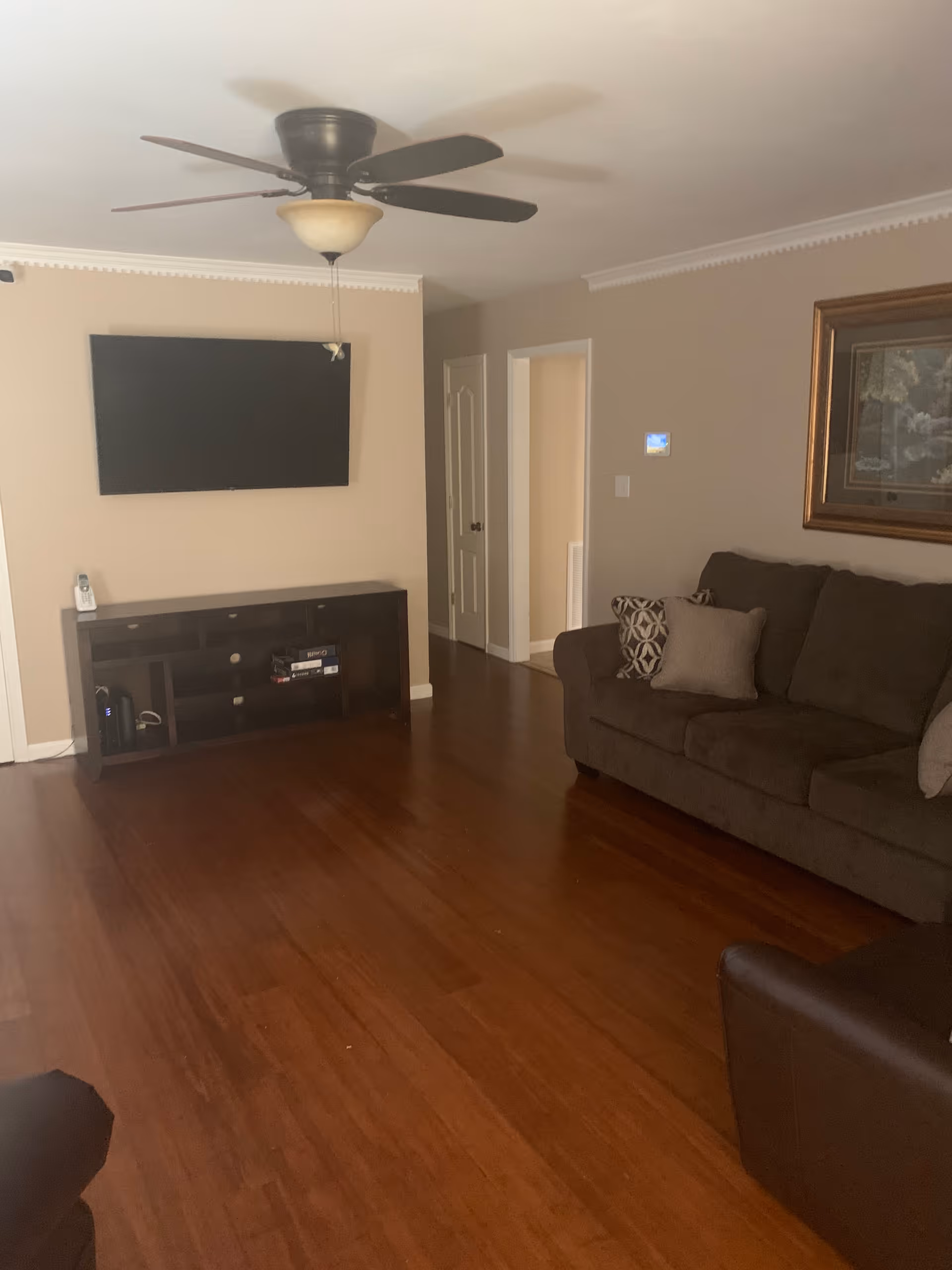 A living room with wooden flooring, a ceiling fan with light, a wall-mounted flat screen TV above a dark wooden TV stand, a brown sofa with patterned and plain cushions, and a framed painting on the wall.