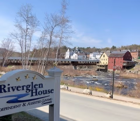 View of a river with a bridge and several buildings in the background under a clear blue sky. In the foreground, there is a white sign that reads 'Riverglen House Independent & Assisted Living Community.'