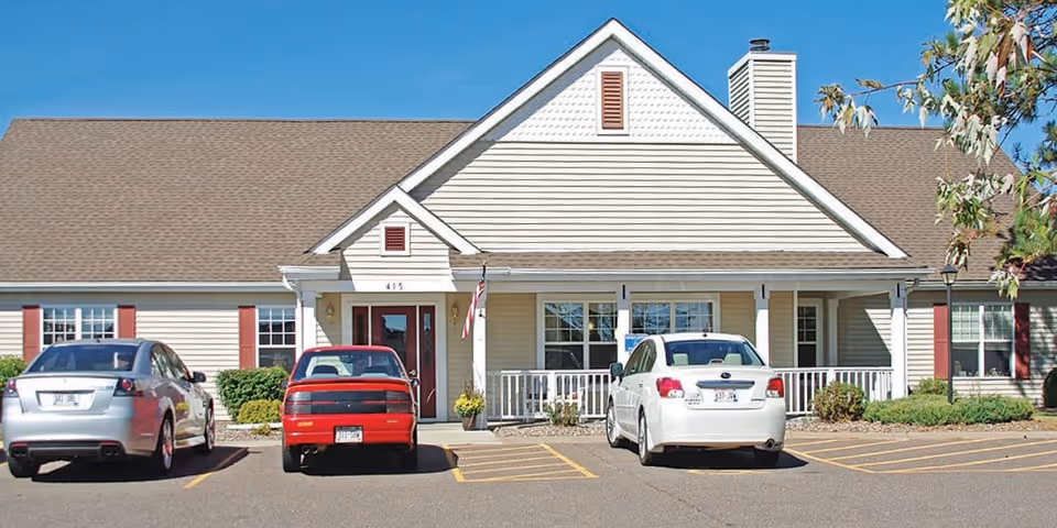 Front exterior view of a single-story senior living facility building with beige siding, a brown shingled roof, and red shutters. Three cars are parked in front of the building in marked parking spaces. There is a small porch with white railings and an American flag near the entrance.
