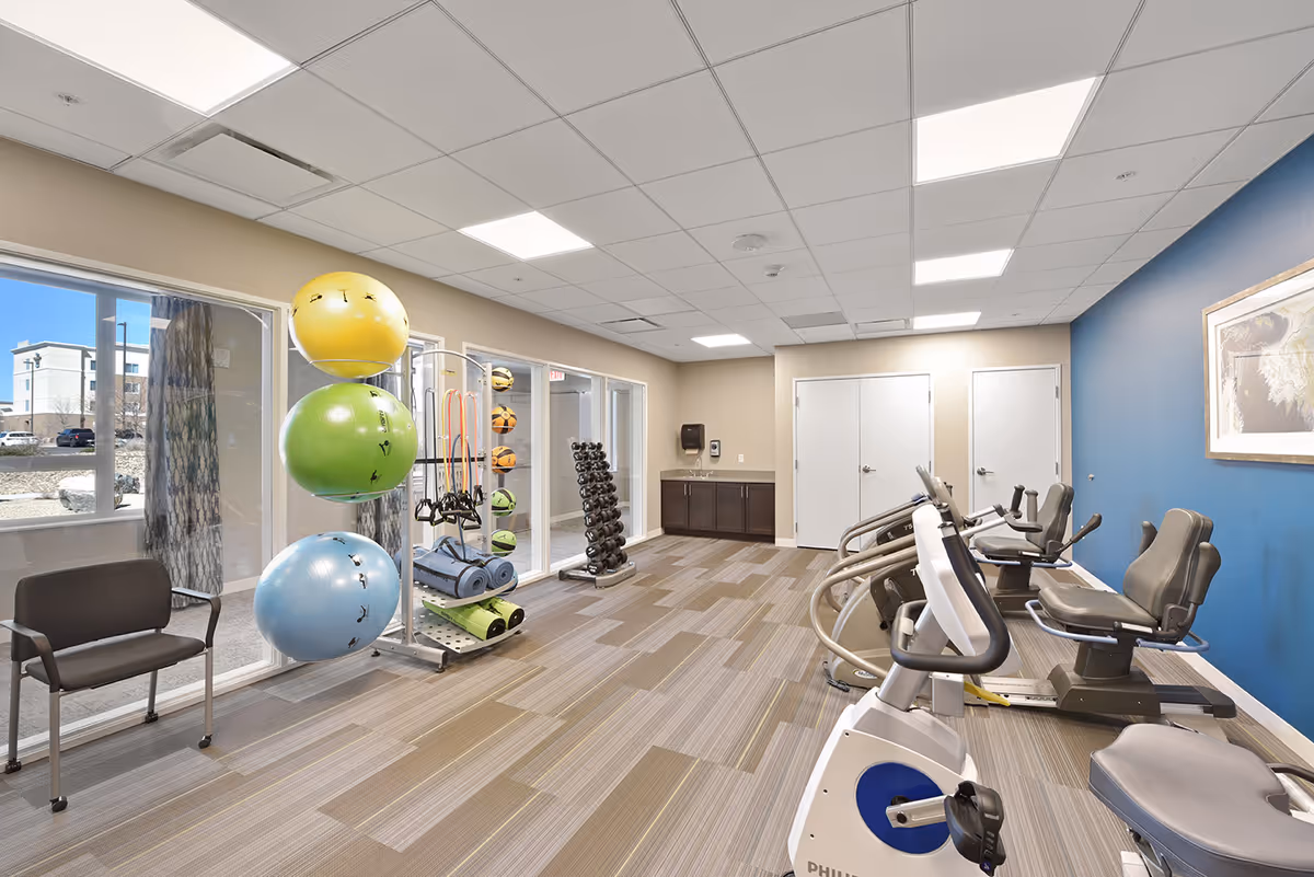 A bright fitness room with exercise equipment including stationary bikes, a rack of dumbbells, large colorful exercise balls, yoga mats, and resistance bands. The room has large windows letting in natural light, beige walls with one blue accent wall, and a chair near the window.