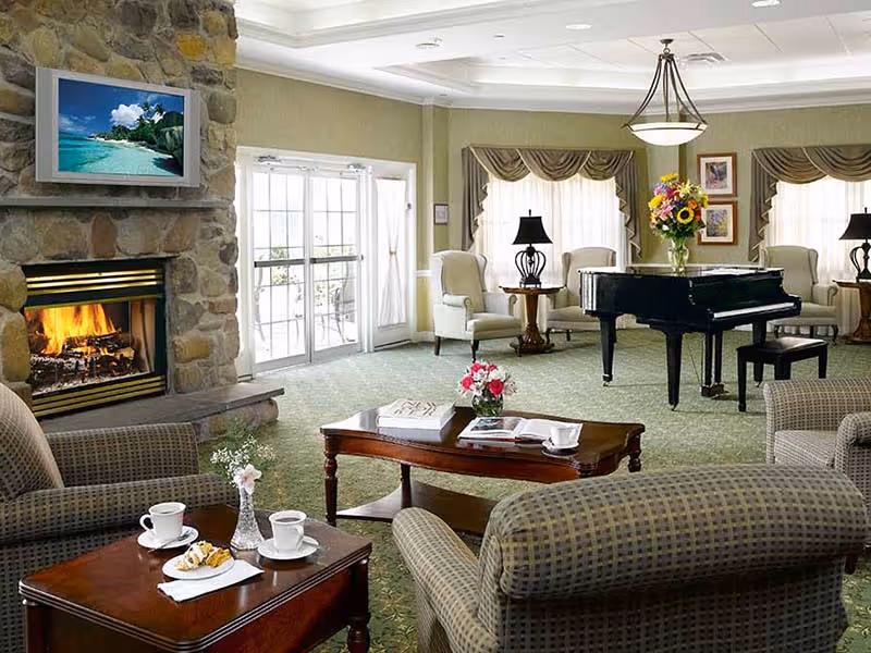 A cozy living room area with a stone fireplace, a flat-screen TV mounted above it, and a grand piano in the center. The room features comfortable armchairs and sofas, a wooden coffee table with flowers and books, and large windows with draped curtains allowing natural light to fill the space.