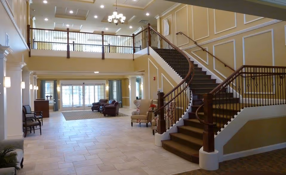 Spacious and well-lit interior lobby area of a senior living facility with a large staircase featuring wooden handrails and carpeted steps. The space includes seating areas with armchairs, tiled flooring, and large windows allowing natural light to enter. The walls are painted in a warm beige tone with white trim and decorative molding, and a chandelier hangs from the ceiling.