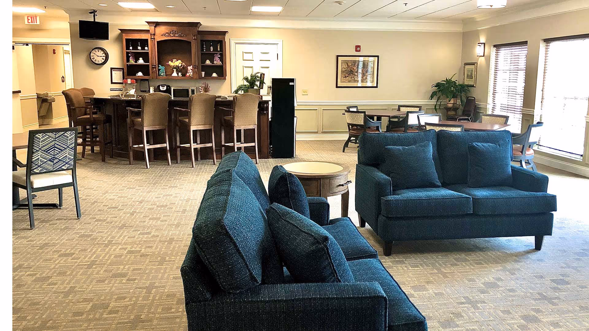 A spacious common area in a senior living facility featuring two blue upholstered sofas with cushions, a round wooden side table, a bar counter with several high chairs, and multiple dining tables with chairs near large windows with blinds. The room is well-lit with ceiling lights and has beige walls and carpeted flooring. There are decorative plants and framed artwork on the walls.
