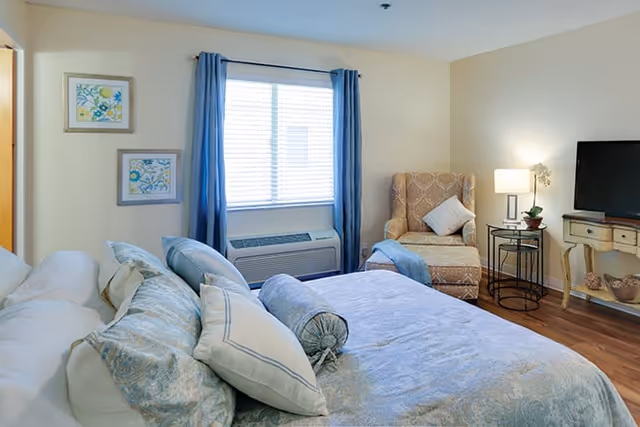 Sunlit bedroom with a made bed, an upholstered armchair, TV on a console, and a window with blue curtains.