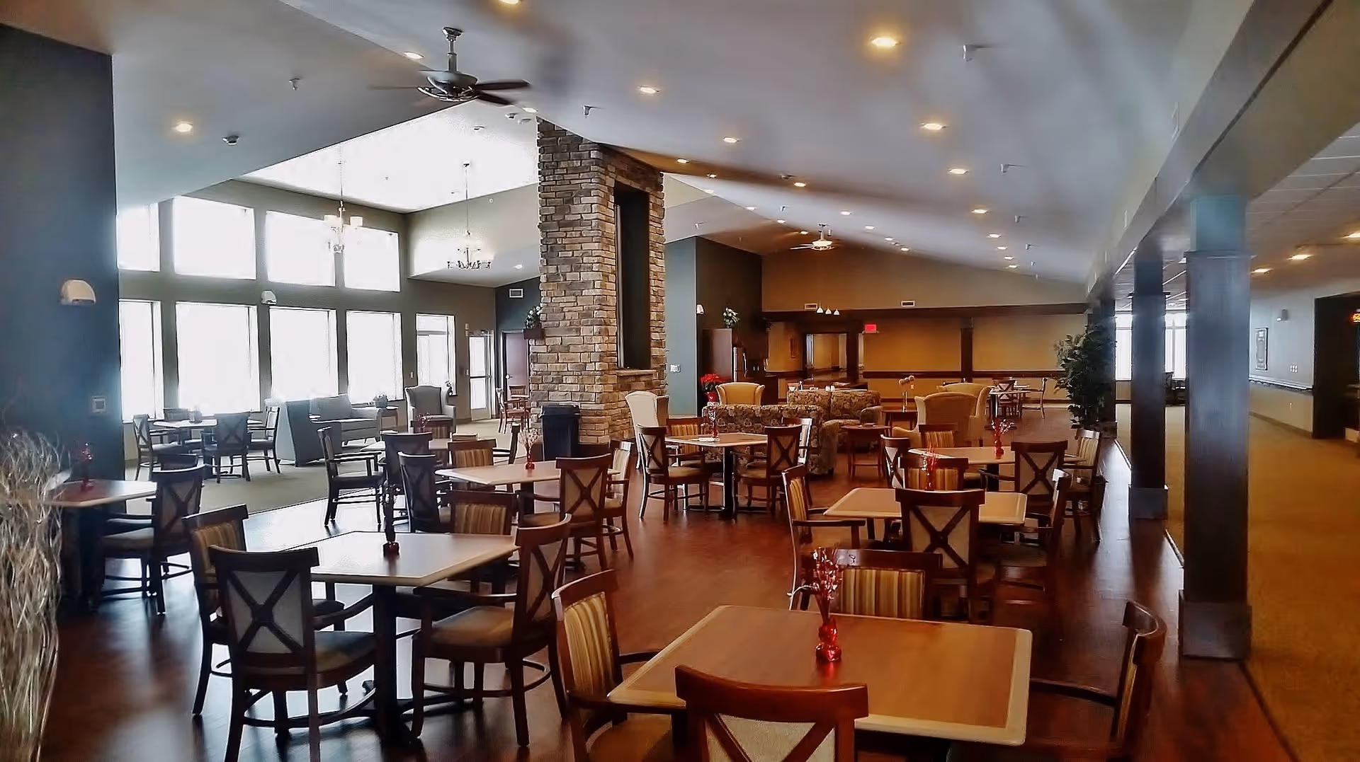 Spacious dining area in Frontida Assisted Living of Elkhorn with multiple wooden tables and chairs arranged neatly. The room features a high ceiling with recessed lighting and ceiling fans, large windows allowing natural light, a stone pillar in the center, and comfortable seating areas with sofas and armchairs in the background.