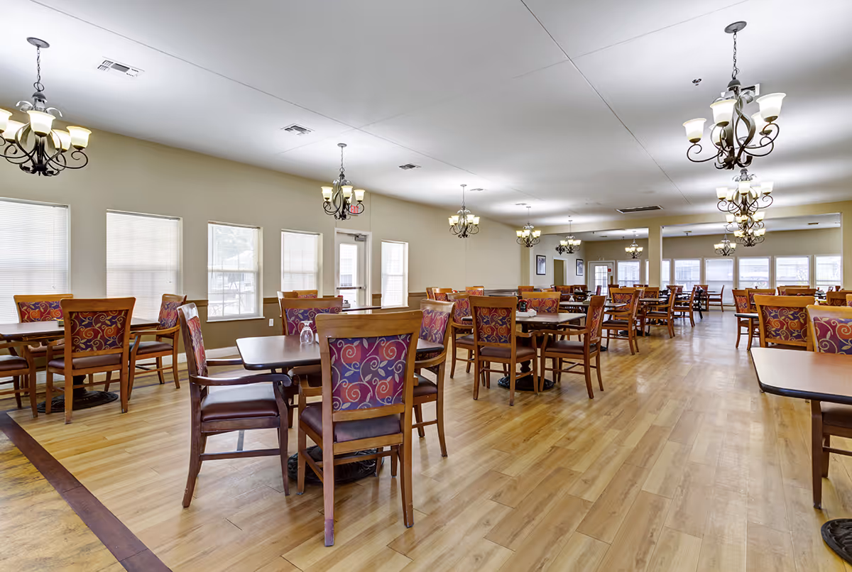 A spacious dining room with multiple wooden tables and chairs featuring patterned upholstery. The room has large windows with blinds, light-colored walls, wooden flooring, and several ornate chandeliers hanging from the ceiling providing warm lighting.