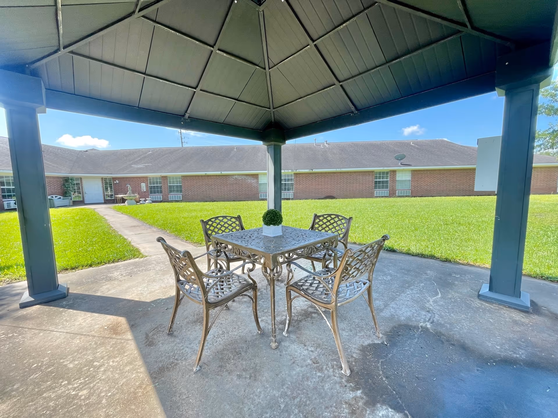 Outdoor covered patio area with a metal table and four matching chairs on a concrete floor. The patio overlooks a grassy courtyard with a brick building in the background under a clear blue sky.