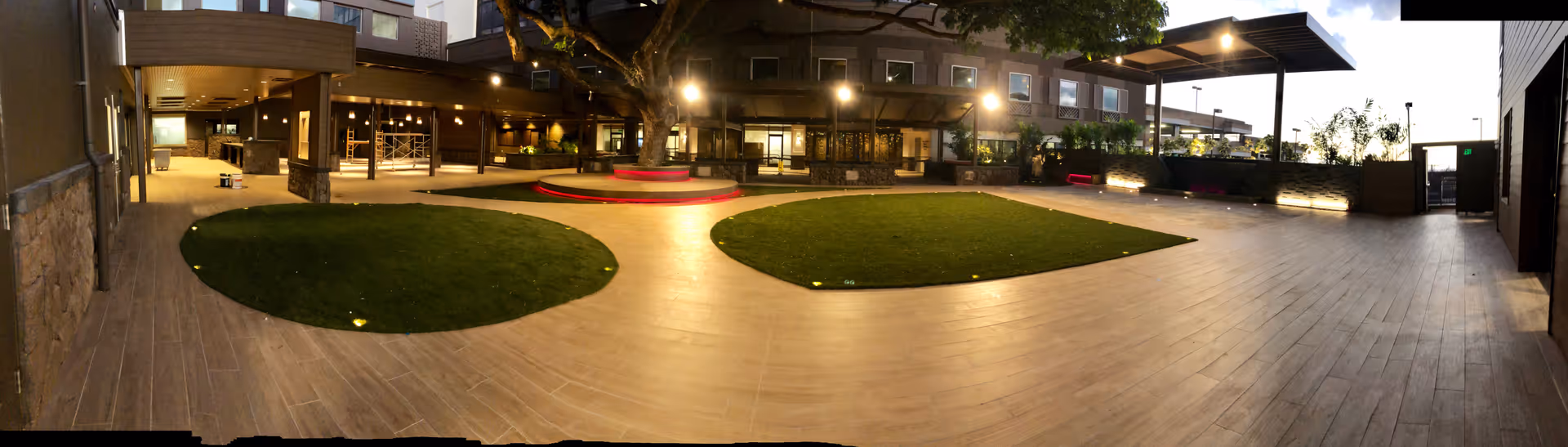Outdoor courtyard area at dusk with two circular grass patches surrounded by wooden flooring, modern building with large windows and balconies in the background, and overhead lighting illuminating the space.