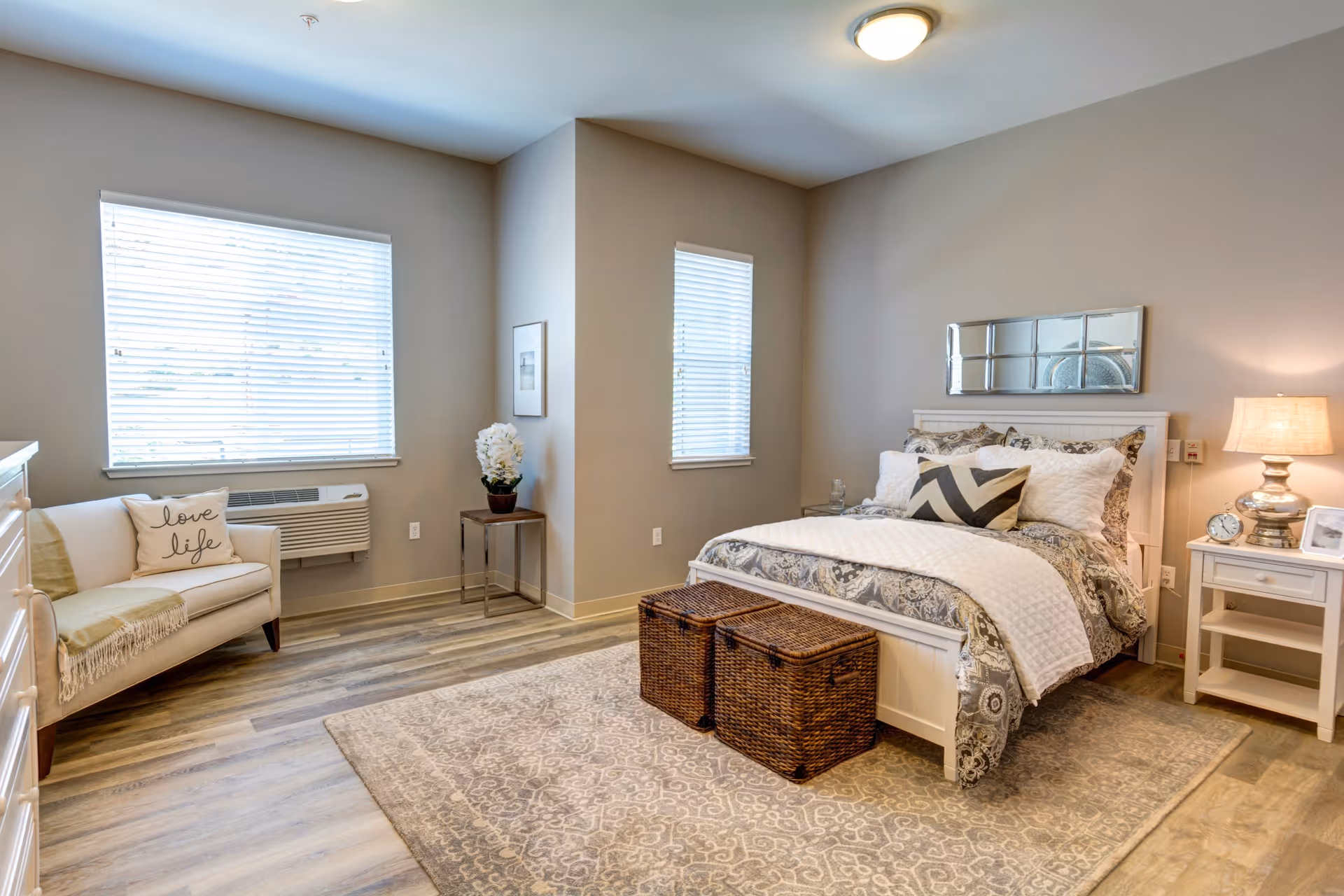 A cozy bedroom with a large bed featuring patterned bedding and multiple pillows. Two woven storage baskets are placed at the foot of the bed. A nightstand with a lamp, clock, and picture frame is on one side of the bed. There is a small white loveseat with a pillow that says 'love life' near a window with blinds. The room has light-colored walls, wood flooring, and a decorative rug.