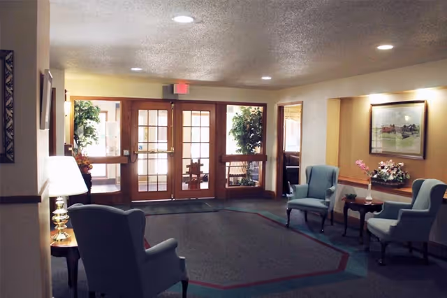 A cozy interior common area with four blue upholstered armchairs arranged around two small wooden tables with floral arrangements. The room has beige walls, a framed picture on the wall, and large glass double doors leading outside. There are potted plants near the entrance and ceiling lights providing illumination.